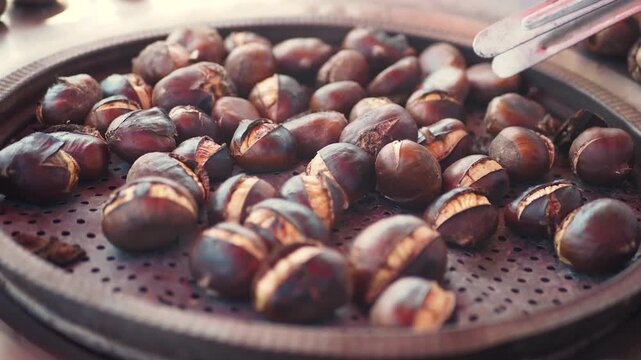 Roasted chestnuts being turned with tongs on a large perforated cooking surface, close-up shot of hot street food being prepared