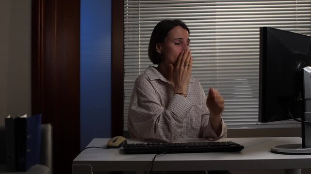 Businesswoman feeling exhausted and sleepy, covering her mouth while yawning during a long night shift at the computer in a dimly lit office environment, depicting burnout and overtime