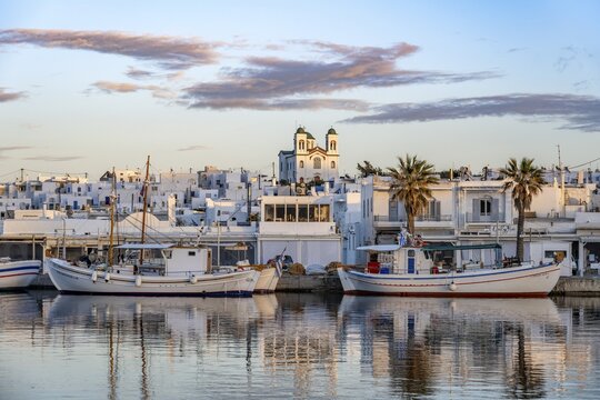 View of Naoussa, Fishing boats in the harbour at sunset, reflected in the sea, White Cycladic houses and church Agios Faneromeni, Naoussa, Paros, Cyclades, Greece