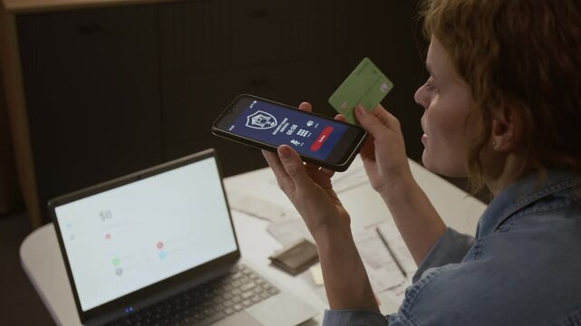 Medium rear shot of distressed Caucasian woman holding smartphone on loudspeaker and credit card, talking to fake bank security service, revealing credentials while watching money stolen from account