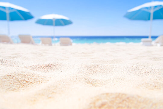 Close-up of clean white sand in the foreground with blurred blue sun umbrellas and lounge chairs on a sunny beach background. Concept of summer vacation and luxury resort relaxation