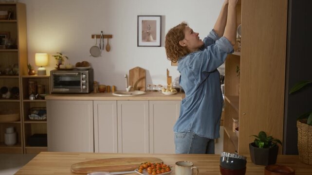Medium shot of middle-aged woman removing plates and bowls from cupboard while making lunch in kitchen, experiencing sharp pain after reaching for top shelf, massaging shoulder and grimacing