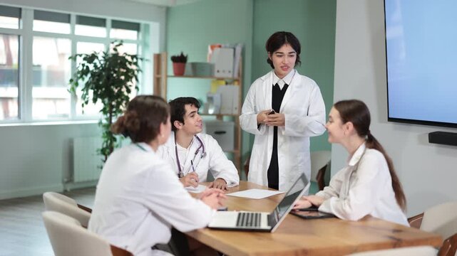Young female doctor in medical gown holds meeting at table with group of colleagues