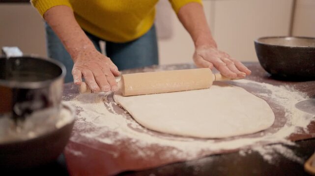 Hands rolling dough with wooden pin on floured table. Shaping pastry base. Bowl nearby holding remaining mixture. Baking process in close detail. Focusing on movement and preparation during cooking.