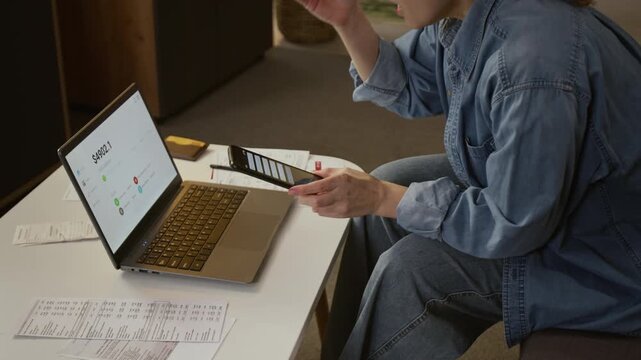 Medium arc shot of mature Caucasian female scan victim watching money disappear from bank account on laptop, grabbing phone with alerts in panic, while dealing with personal finances at home