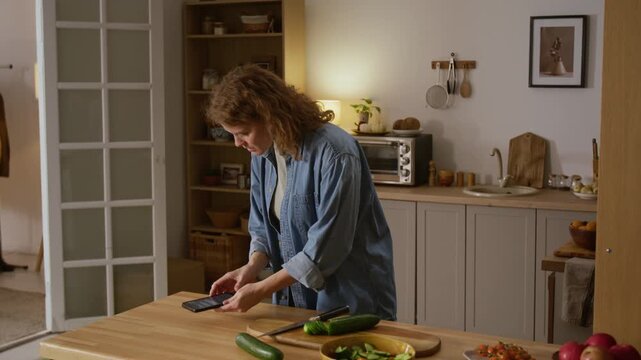 Medium full shot of Caucasian woman cutting vegetables for lunch in kitchen, seeing messages of money transferred from bank account on smartphone, then calling support with fear and stress on face