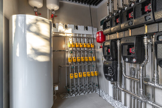 In a utility room, pipes and tanks are neatly organized on the wall. Gauges and control systems are placed for operation. Light from outside shines on the setup