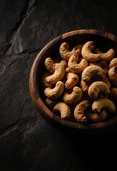 Fototapeta premium Golden roasted cashew nuts served in a rustic wooden bowl on a dark stone background, healthy and crunchy snack