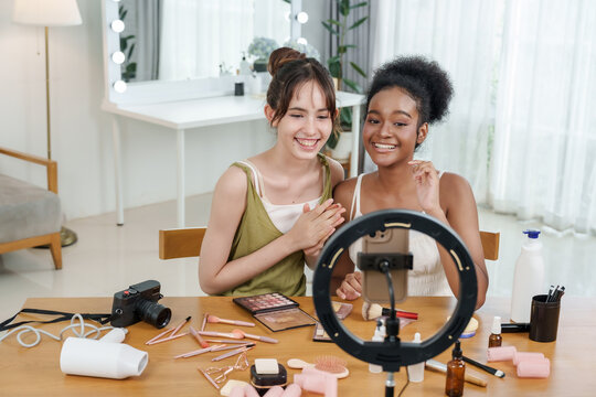 asian teenage content creator filming video with black teenage friend smiling while preparing makeup tools for skincare vlog tutorial concept beauty influencer cosmetics fun diverse friendship shoot