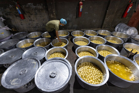 Bogura, Bangladesh - 17 April 2022: View of a worker preparing a vast array of shiny, golden-syruped rasgullas in large metal pots, reflecting light in a dimly lit room.
