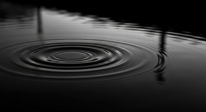 Concentric water ripples spread on a reflective surface in a dark monochrome close-up against a pure black background.