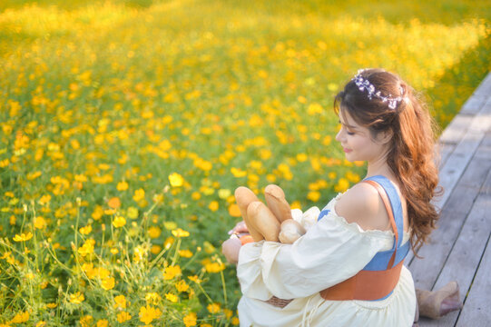 Side view of a Young woman in a romantic cottagecore Renaissance type dress sitting on a wooden deck by a field of yellow flowers holding a basket of baguettes, Thailand