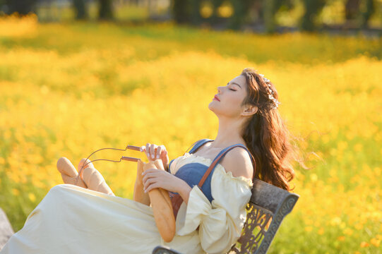 Side view of a Young woman in a romantic cottagecore Renaissance type dress sitting in a chair in a field of yellow flowers holding a baguette, Thailand