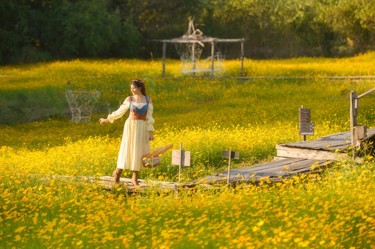 Young woman in a romantic cottagecore Renaissance type dress standing in a field of yellow flowers carrying a basket of baguettes, Thailand