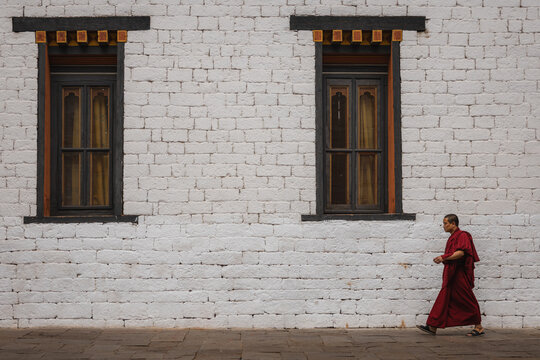 Thimphu, Bhutan - 28 September 2025: View of a lone monk in vibrant saffron robes strides along a whitewashed brick wall adorned with dark, ornate window frames, a study in contrast.