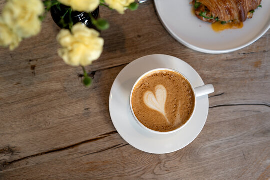 Overhead view of a cappuccino with a heart shaped decoration and a fried egg croissant with greens on a wooden table with flowers in a vase