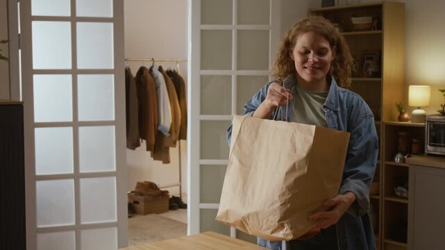 Medium shot of mature Caucasian woman walking through kitchen door, carrying heavy bag with groceries, putting on table then grabbing on to shoulder and spilling purchases after feeling sharp pain