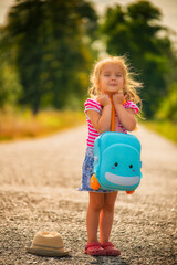little blonde girl standing on a rural road holding a turquoise plastic suitcase, eyes closed in anticipation and joy. straw hat nearby, warm summer light, carefree travel and childhood concept.
