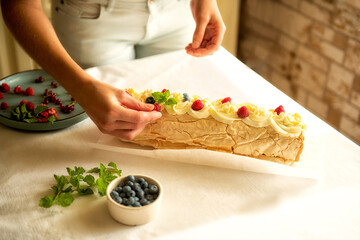 process of making homemade meringue roll cake decorated with fresh berries and mint. female hands adding raspberries on top, cozy kitchen atmosphere, baking at home concept.