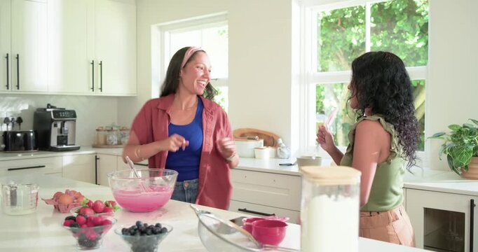 Indian and African American friends stirring pink batter at kitchen island checking phone dancing