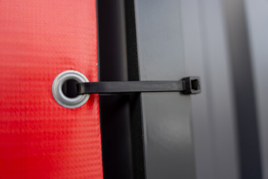 Close up of black strap fastener through metal grommet on red textured fabric panel, modern industrial securing mechanism detail with gray background