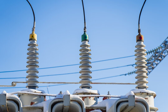 Three high voltage substation insulators with colored phase markers and overhead cables, snow on equipment and clear blue sky background, power grid safety theme