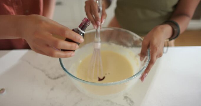 Indian and African American women tipping bottle and whisking in bowl on counter, coloring batter