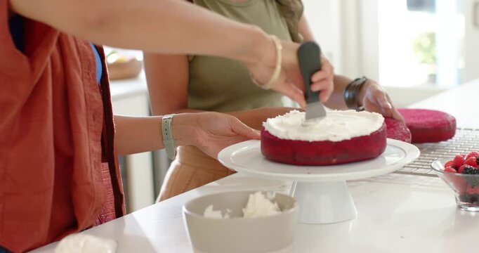 Female friends frosting 2-layer red cake in kitchen using spatula one holding stand forming layers