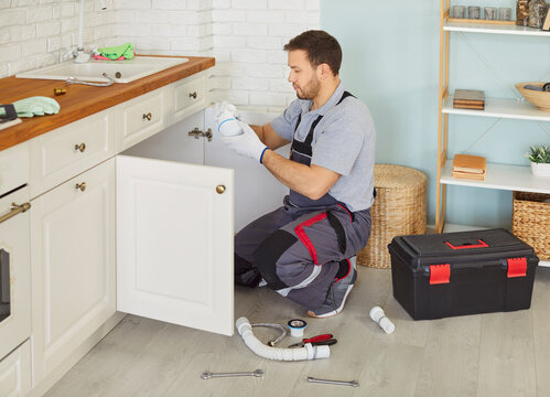 Professional male plumber in uniform with toolbox sitting near sink replacing the water filter or fixing broken pipe in modern kitchen. Repair service and plumbing maintenance concept.