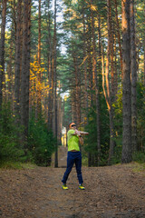 Obraz premium Man in activewear stretching arms outdoors on a wide forest path surrounded by tall pine trees, engaging in physical activity and promoting a healthy lifestyle in nature