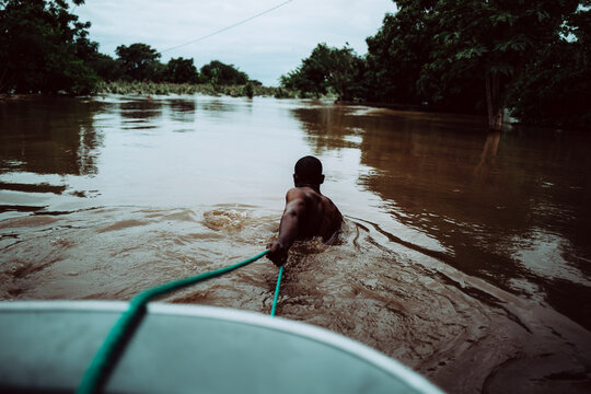 Manhica, Mozambique - 20 January 2026: View of a man waist-deep in the murky, swirling floodwaters, pulling a boat forward through the submerged landscape.