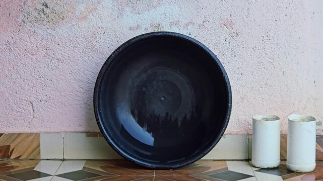 After washing clothes, a wet black tub leans against a wall to dry on tiles. Beside it, two repurposed white bottles, cut into cups and filled with water, sit ready for further household utility tasks