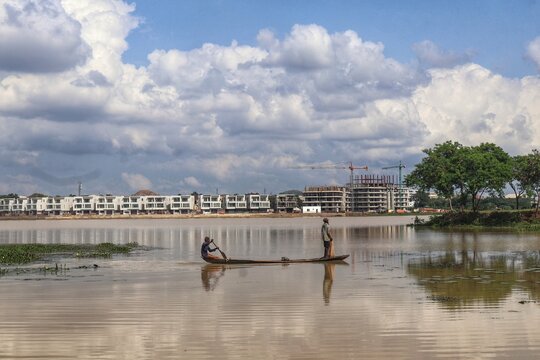 Abuja, Nigeria - 17 August 2024: View of a serene waterway reflects the sky, with a slender canoe carrying two figures gliding past modern buildings near Alex Ekwueme Way.