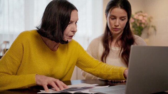 Experienced Caucasian woman pressing laptop keys at table. Beautiful female guiding and watching screen attentively. Papers lying nearby during lesson. Learning digital skills at home kitchen.
