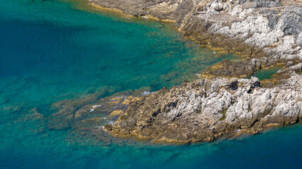 Crystal-clear turquoise water gently laps against a rugged, light-colored rocky shoreline. The varying depths create a stunning gradient of blue and emerald tones.