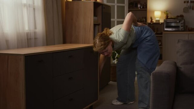 Medium full shot of young Caucasian woman trying to move cabinet drawers while tidying up at home, then crying from sudden back injury, standing up and rubbing muscles around waist area