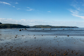 Low tide landscapes along the beaches of Baie Sainte Catherine with sandy and pebbled shoreline, calm shallow water, scattered rocks and forested hills in the background under clear blue sky