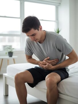 Young man sitting on bed clutching stomach in pain indoors