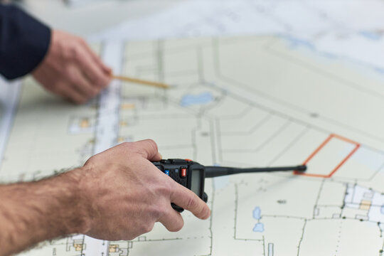 Middle aged Caucasian man holding walkie talkie and pointing pencil at architectural blueprint, working in office environment, demonstrating foreman responsibilities and coordination