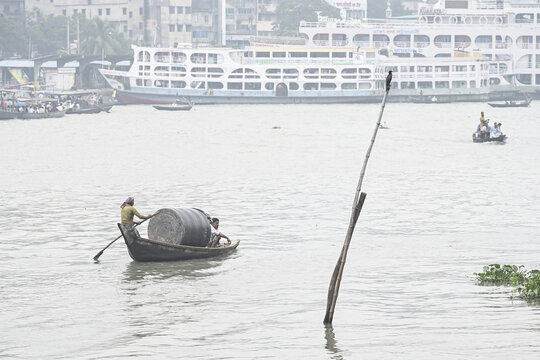 Dhaka, Bangladesh - 09 September 2025: View of the murky river reflecting the overcast sky as a small boat laden with cargo is rowed past anchored ferries.