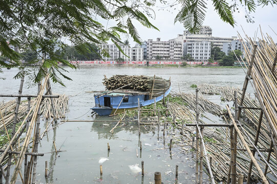 Dhaka, Bangladesh - 10 September 2025: View of a weathered blue boat laden with timber floating on the murky water, framed by bamboo structures, against a backdrop of urban buildings.