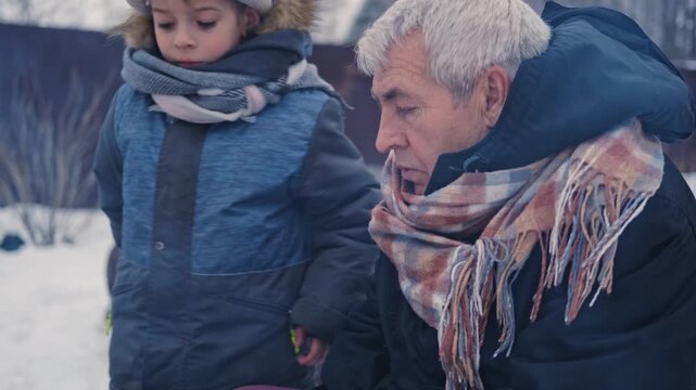 Gray haired grandfather and child by grill roasting sausages on charcoal brazier, bundled in winter coats and scarves on snowy ground. Hands warmed over glowing embers, smoke. Family outdoor barbecue.