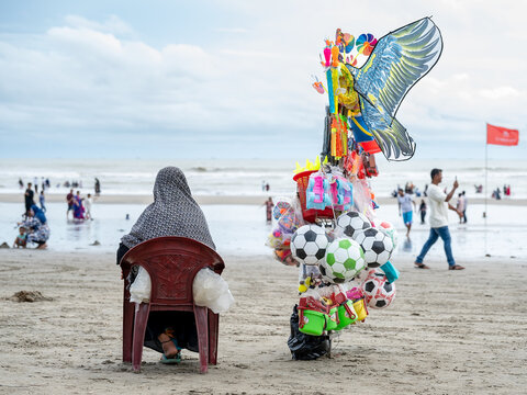 Cox's Bazar, Bangladesh - 08 August 2025: View of a beach vendor seated near colorful kites and inflatable balls against a vast, soft sandy beach, with the gray ocean stretching to the horizon.