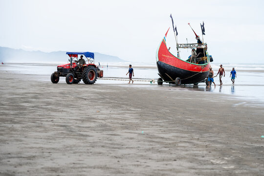 Cox's Bazar, Bangladesh - 09 August 2025: View of a vibrant red and black fishing boat being pulled onto the sandy beach by a red tractor under a cloudy sky.