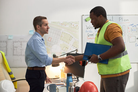Caucasian middle aged man standing at desk shaking hands with Black middle aged man wearing safety vest, holding clipboard in office with construction plans on wall