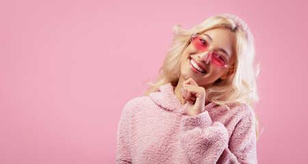 A young woman stands with a playful smile while wearing pink sunglasses. She has long, light-colored hair and is dressed in a cozy, pink sweater. The background is a solid pink color. © Prostock-studio