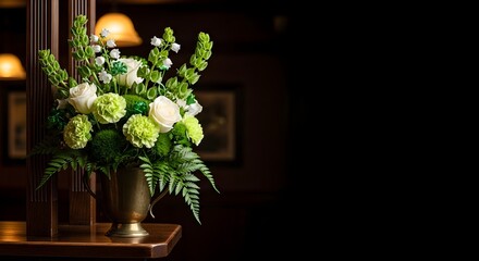 Elegant floral arrangement featuring white roses, green carnations, and lush ferns in a brass vase, set against a dark, softly lit background with warm lamp glow.