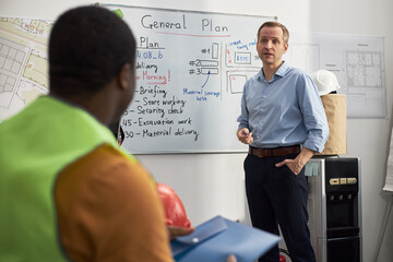 Caucasian middle aged man explaining project plan on whiteboard to Black man in office setting,...