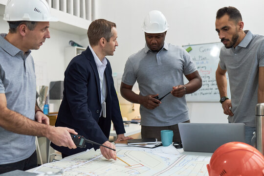 Middle aged Caucasian man in suit discussing construction plans with two middle aged men and one young adult Black man wearing hard hats in office, all standing around table with blueprints