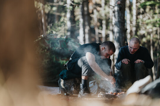 Two men sit by a campfire in a wooded area. They relax and chat as smoke rises in a calm outdoor campsite.